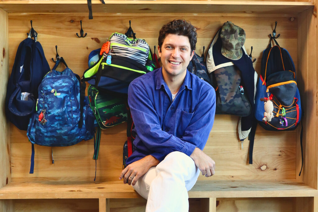 Colton Eddy | STEAM School Lead Educator A man with curly brown hair, wearing a blue chore jacket and white pants, sits smiling on a wooden bench in a mudroom-style cubby unit. Rows of children's backpacks, lunch bags, and a camo baseball cap hang from hooks on either side of him.