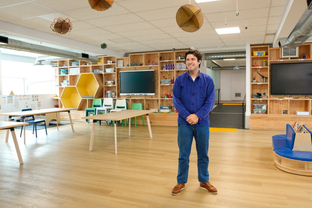 Colton Eddy | STEAM School Lead Educator A man in a blue chore jacket, jeans, and brown work boots stands smiling in a bright makerspace classroom. Wooden shelving units filled with STEM supplies and bins line the walls, with a large screen mounted at the back, wooden tables and stacked chairs nearby, and geometric wooden pendant lights hanging from the ceiling.