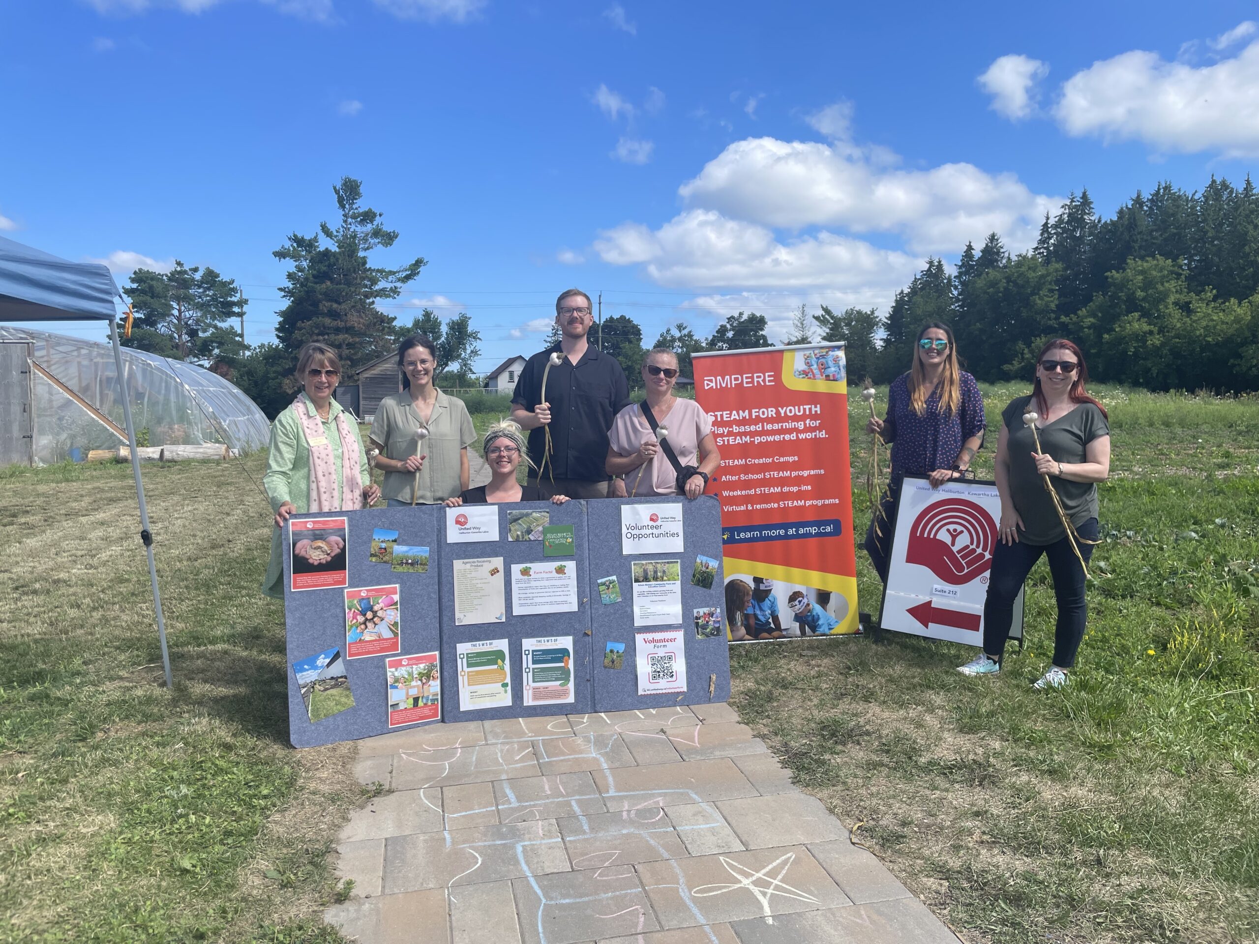 A group consisting of Ampere employees and those connected to OTF and United Way is posed in front of a greenhouse. It's daylight.