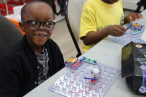 A child sits at a table wearing glasses and a black coat. He is learning with a STEAM activity in front of him. Another child with a yellow shirt is beside him.