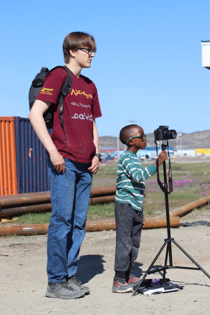An instructor and a child stand behind a camera. They are outside.