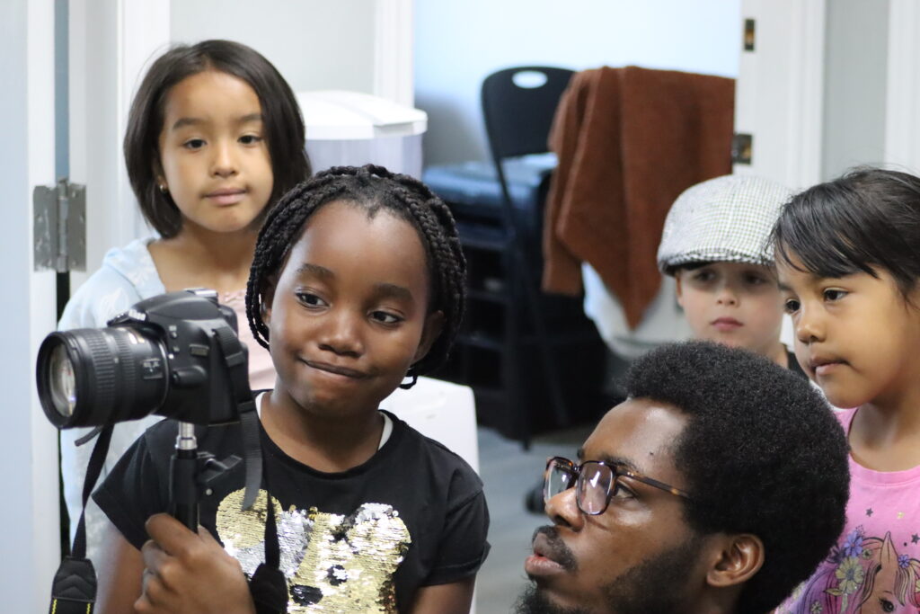 One adult and three campers look at a digital camera pointed to the left. They are indoors.