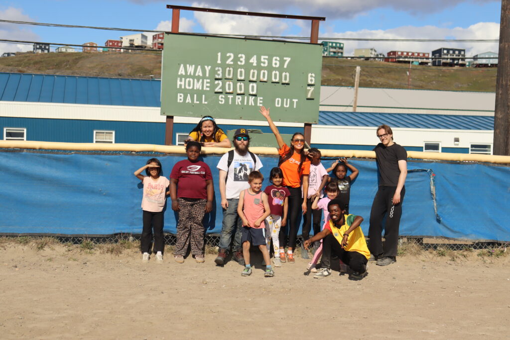 Summer camp attendees stand in front of a score board in a baseball diamond. 