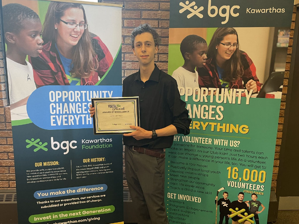 Jamie Jeffrey poses with a BGC award. He is wearing a black shirt and standing in front of two BGC pull-up banners.