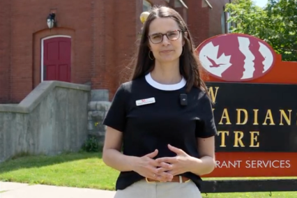 Katelyn stands in front of a sign that reads "New Canadians Centre". Behind her is a large red church.