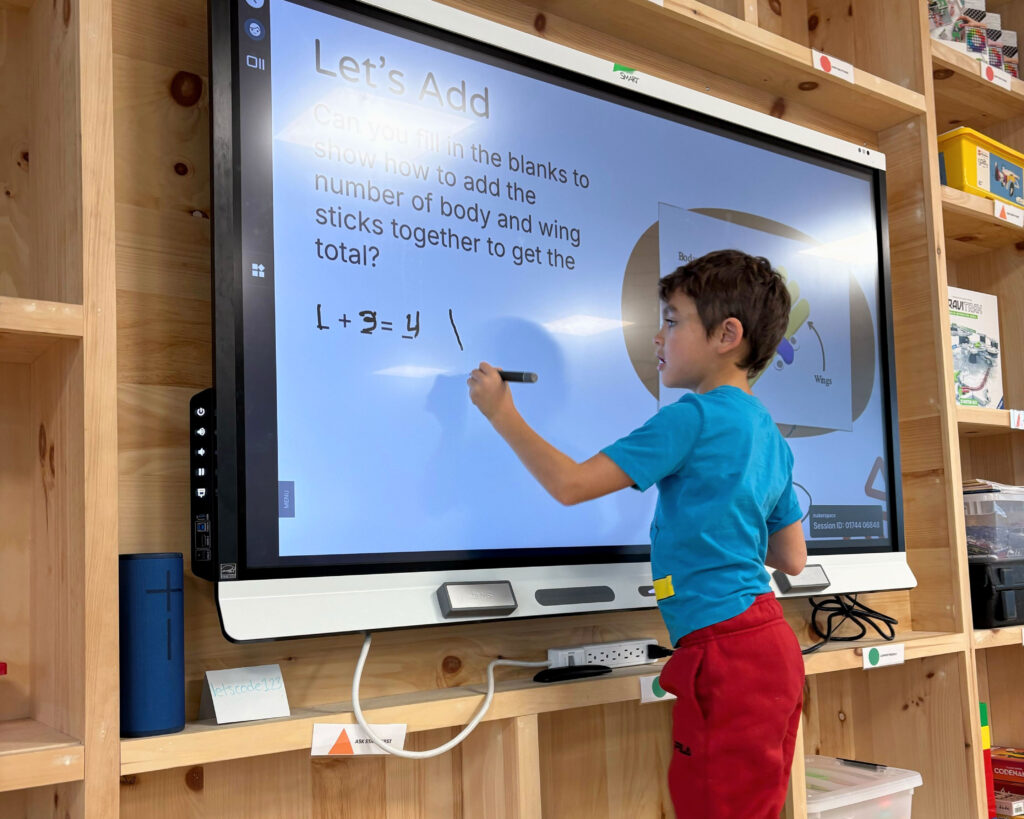 A young boy in a teal shirt and red pants writes on an interactive smartboard displaying an addition exercise titled 'Let's Add,' with a diagram about counting body and wing sticks in the background.