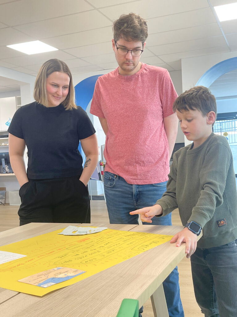 A woman, a man wearing glasses, and a boy stand around a table looking down at a large yellow poster covered in handwritten notes and diagrams, with the boy pointing at the paper.