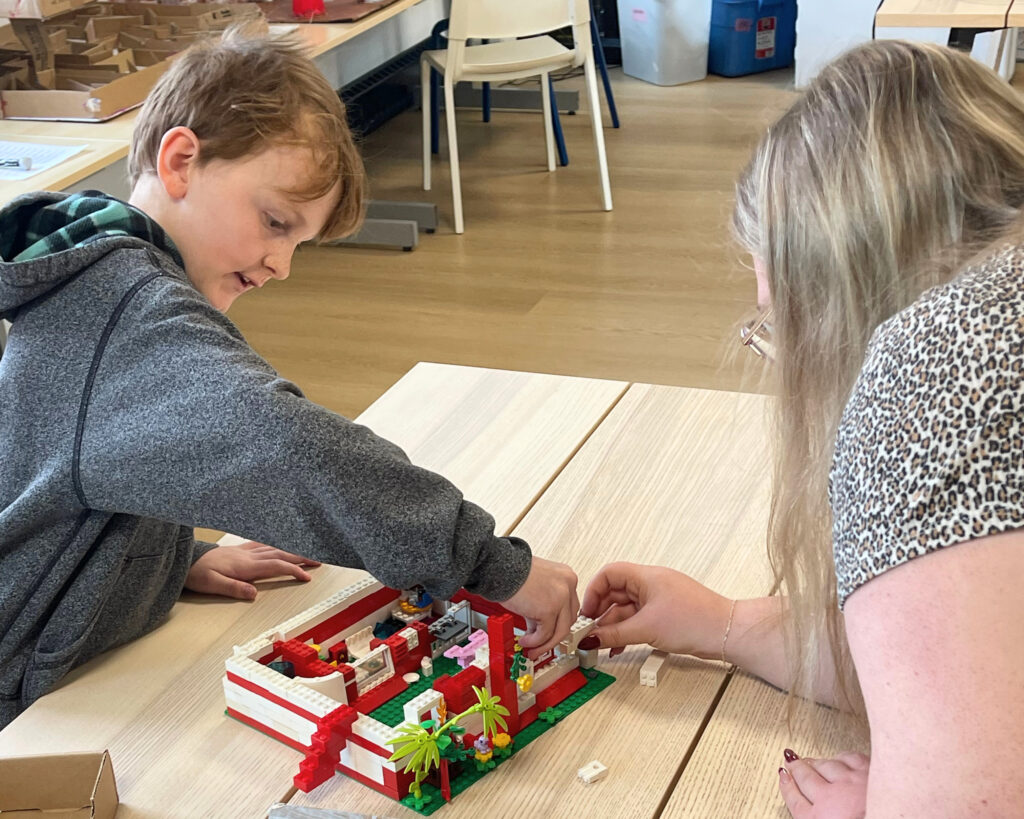 A young boy and a woman work together building a red, white, and green LEGO structure with palm trees on a wooden table, both reaching in to place pieces.