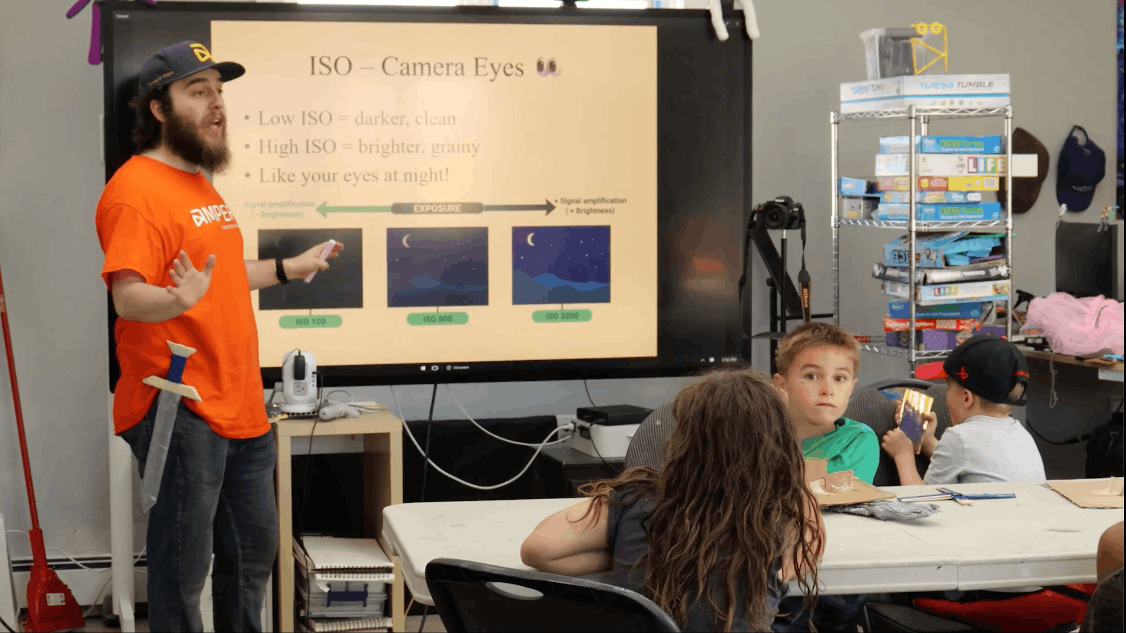 Declan, wearing an orange Ampere t-shirt and cap, stands beside a wall-mounted screen showing a slide titled "ISO – Camera Eyes," and gestures toward three night-sky images labelled ISO 100, ISO 800 and ISO 3200. A cardboard sword hangs from his belt. In the foreground, children sit at a table with cardboard craft pieces, and one boy in a green shirt looks back toward the camera.