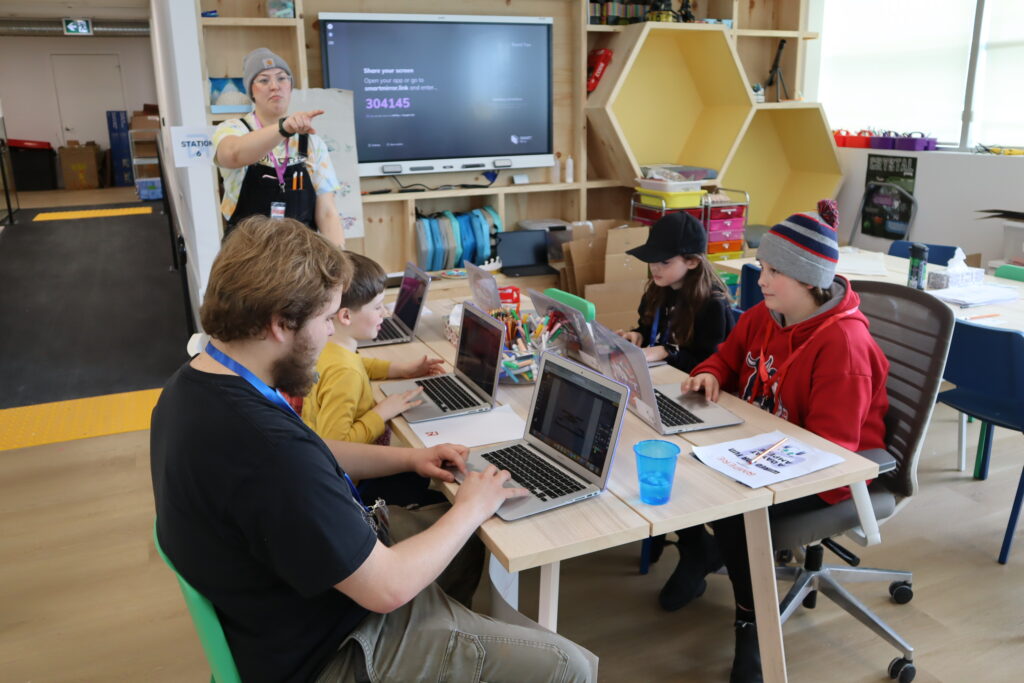 Hakathon Kids working on laptops at a Makerspace table while a facilitator gives instruction.