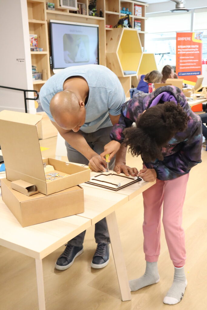 A parent and child are interacting with STEAM activities at a table in the Ampere Kawartha Lakes Makerspace and Skills Hub. A smart board behind them displays STEAM information. They are working on a project called Solar Mindset.