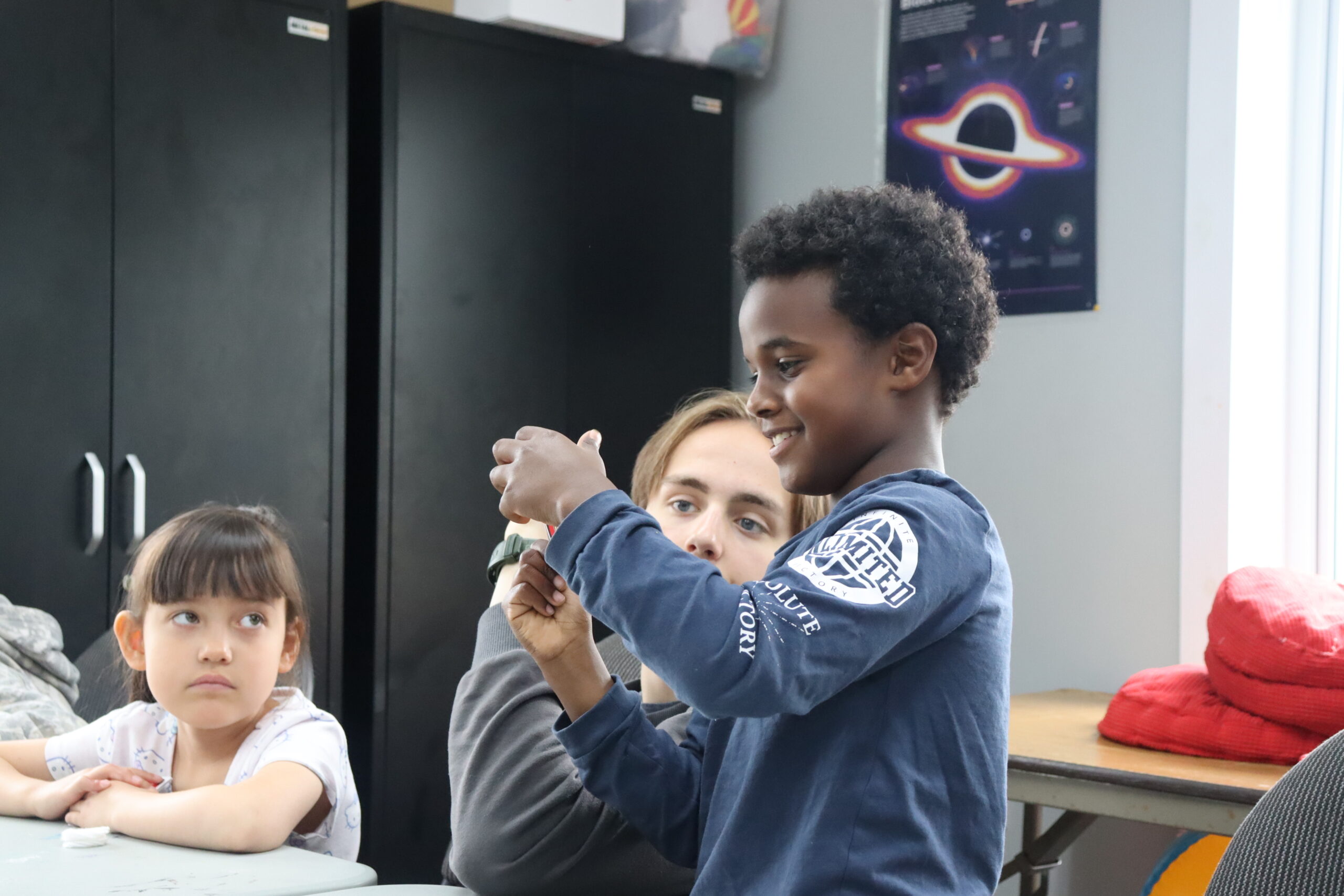 A camper at Ampere's Iqaluit STEAM Summer Camp smiles while examining a wearable device on his wrist, with an instructor and another young camper nearby in the Makerspace.