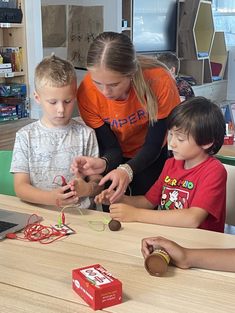 An Ampere instructor helps two campers at Kawartha Lakes STEAM Summer Camp build a Makey Makey circuit out of conductive clay, connected to a laptop on the workbench.