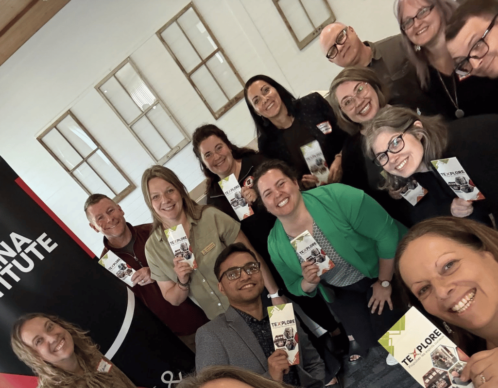 Group selfie of TeXplore Partner Day attendees smiling and holding TeXplore brochures, standing in front of a McKenna Institute banner.