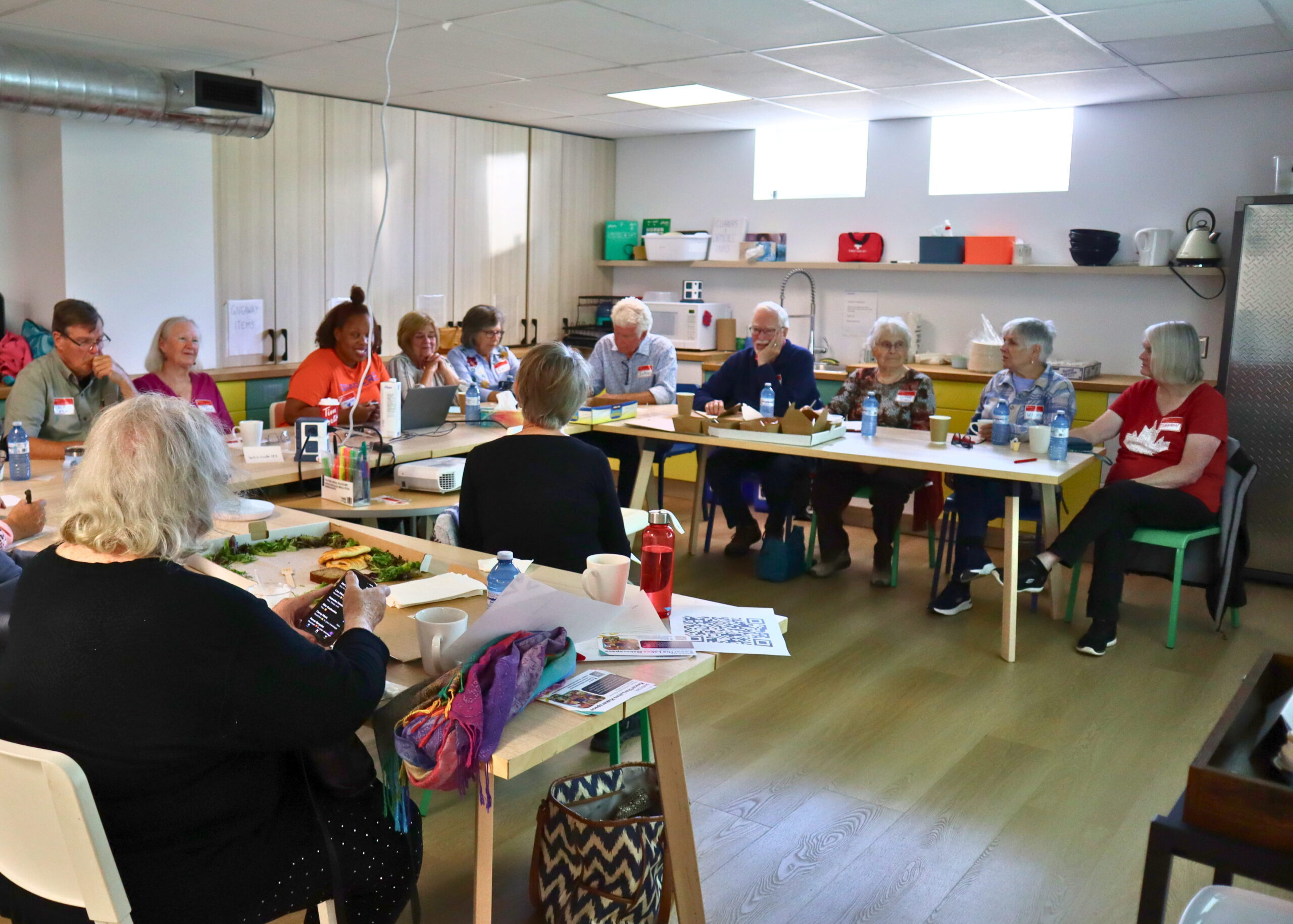 A group of about a dozen older adults, all wearing name tags, sit around a U-shaped arrangement of tables in a bright makerspace with a kitchenette in the background. A woman in an orange t-shirt sits at a laptop and smiles as she speaks to the group, while participants listen. Takeout containers, coffee mugs, water bottles, and printed handouts with a QR code sit on the tables, and one person in the foreground looks at a smartphone.