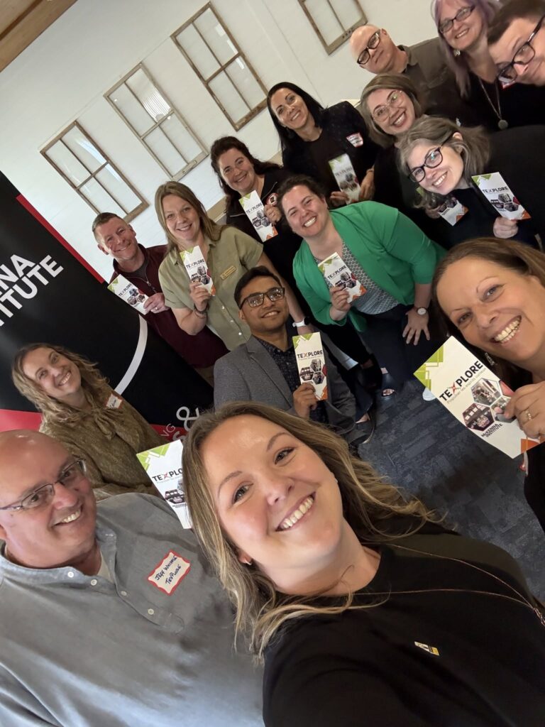 Group selfie of TeXplore Partner Day attendees smiling and holding TeXplore brochures, standing in front of a McKenna Institute banner.
